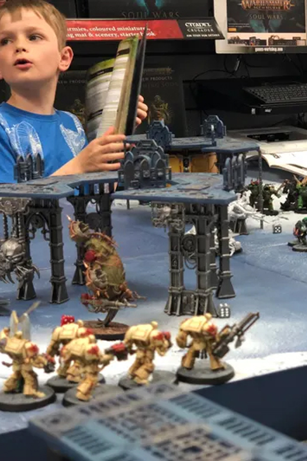 Youth playing miniature wargames with miniatures, table top terrain, and rules books.